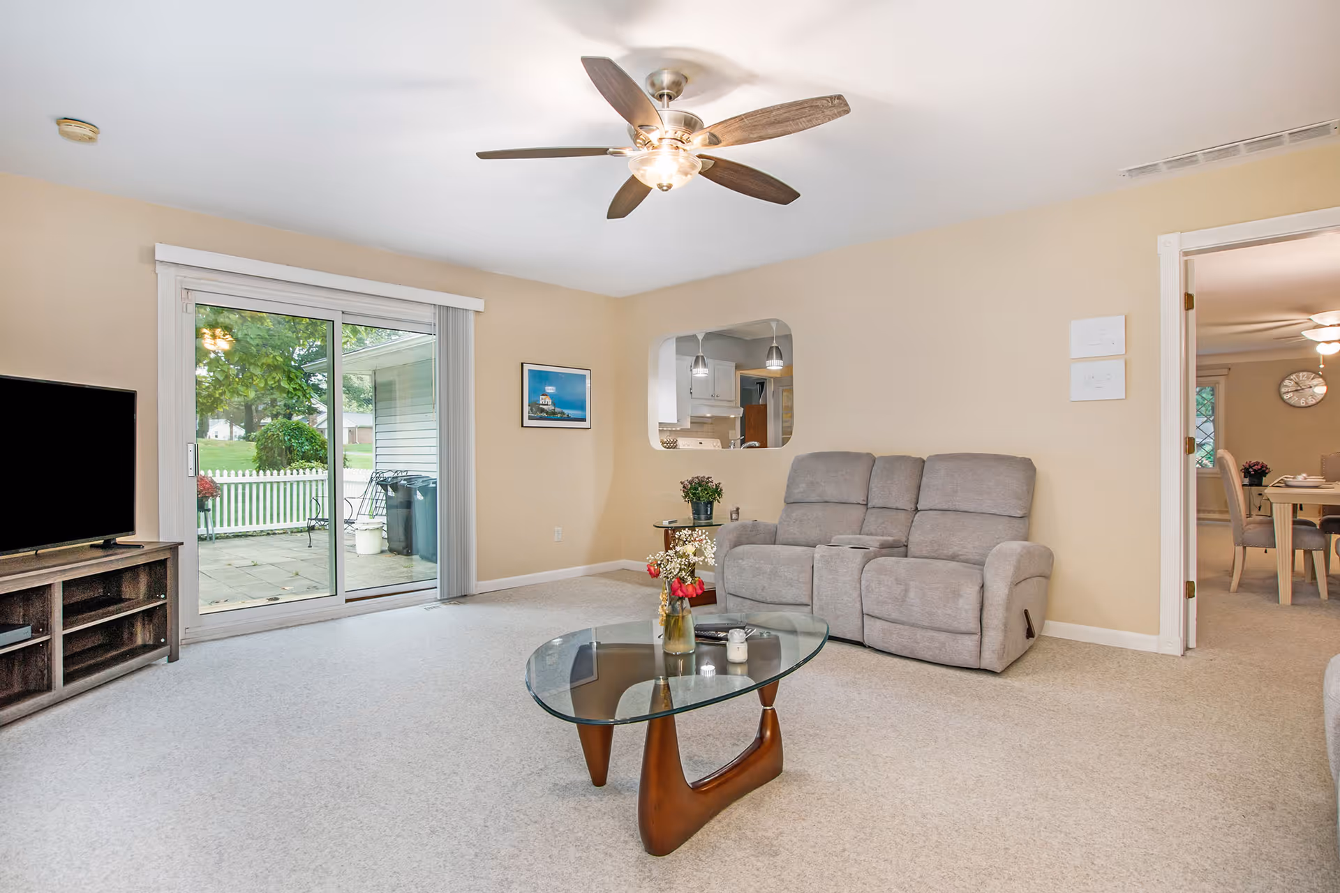 A bright living room with beige walls and carpet, featuring a gray reclining loveseat, a glass coffee table with a wooden base and flower vases on top, a ceiling fan with lights, a flat-screen TV on a wooden stand, and sliding glass doors leading to an outdoor patio with a white picket fence and greenery. A doorway on the right leads to a dining area with a table and chairs.