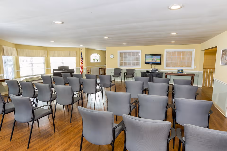 A meeting or activity room with rows of gray chairs arranged facing a wall-mounted television. The room has large windows with blinds, an American flag in the corner, a piano, and light yellow walls with white trim. The floor is wood, and there are additional chairs and tables along the walls.