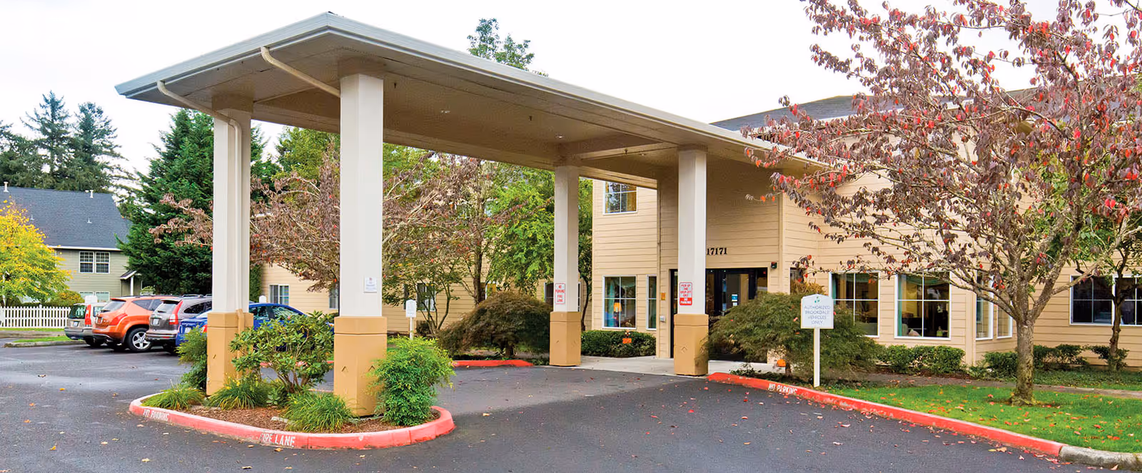 Entrance of a senior living facility with a covered drop-off area supported by four columns. The building is beige with multiple windows and surrounded by landscaped greenery and trees with autumn foliage. Several parked cars are visible to the left.