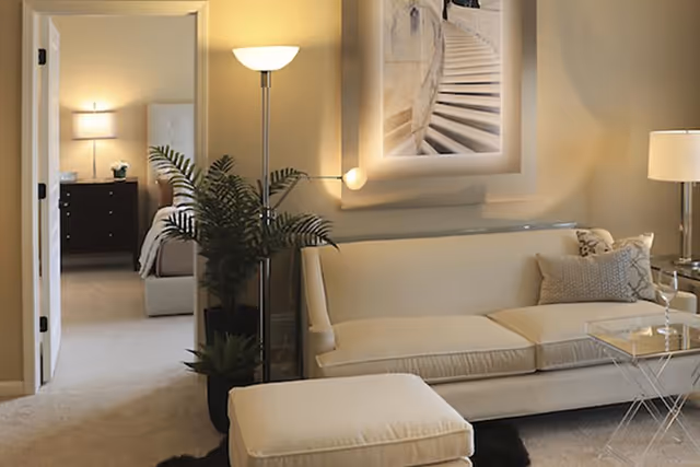 A cozy living room area with a cream-colored sofa, matching ottoman, a floor lamp, and a side table with a lamp. There is a large framed picture of a staircase on the wall. Through an open doorway, a bedroom with a bed, nightstand, and lamp is visible.