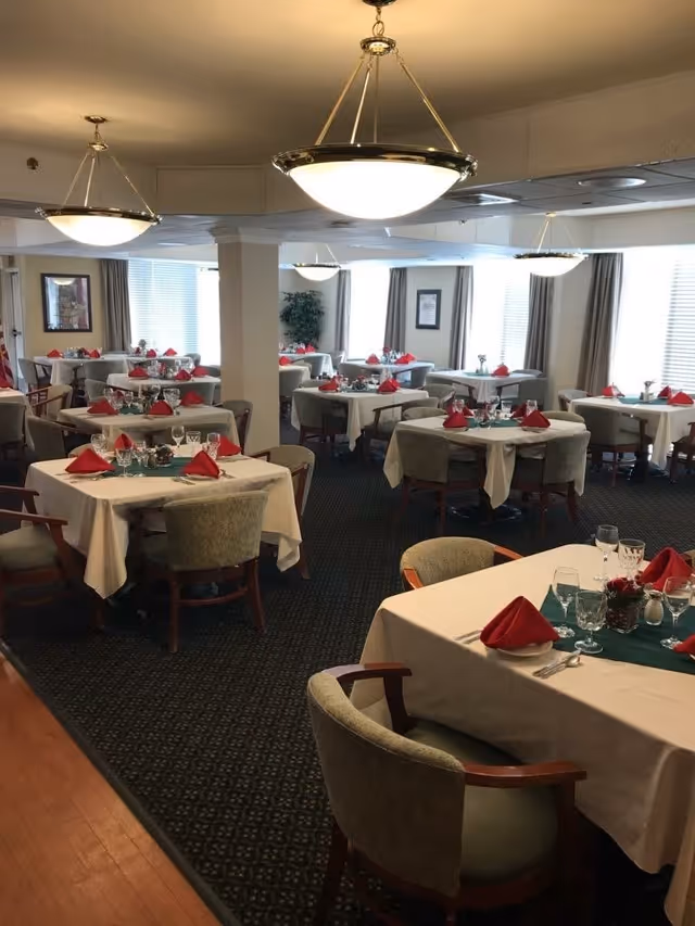A dining room in a senior living facility with multiple tables covered in white tablecloths, each set with red folded napkins, glassware, and silverware. The room has carpeted flooring, large windows with curtains, and ceiling lights hanging from above. Chairs with wooden arms and cushioned seats surround the tables.