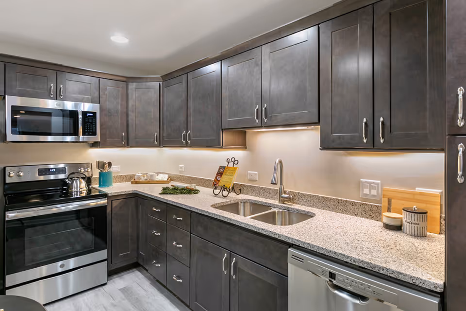 Modern kitchen with dark wood cabinets, granite countertops, a stainless steel stove, microwave, dishwasher, double sink, and various kitchen accessories on the counter.