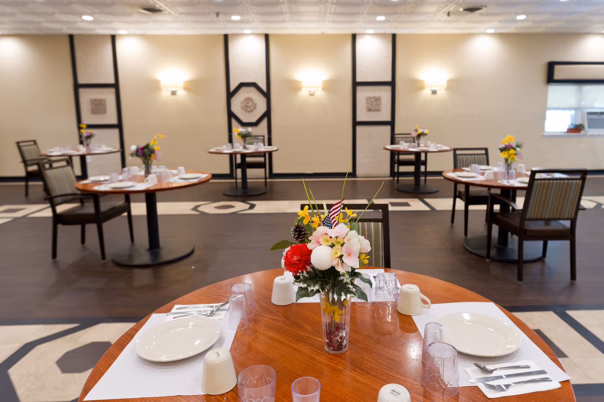 A dining room with round wooden tables set with white plates, cups, utensils, and clear plastic glasses. Each table has a vase with colorful flowers and small American flags. The room has beige walls with decorative panels and wall sconces providing warm lighting. Several chairs are arranged around the tables.