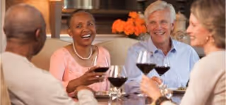 Four adults sitting around a dining table, smiling and holding glasses of red wine, with a vase of orange flowers in the background.
