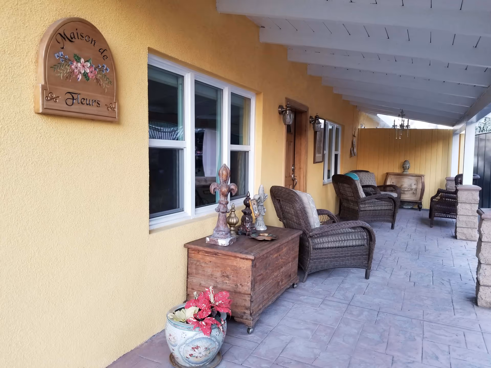 Covered outdoor patio area with yellow walls and white ceiling beams. There are wicker chairs with cushions, a wooden chest with decorative items on top, a potted plant with red flowers, and a wooden sign on the wall that reads 'Maison de Fleurs'.