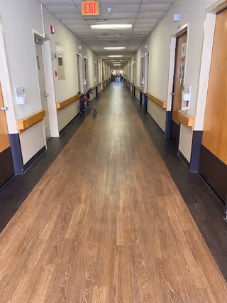 A long, well-lit hallway in a senior living facility with wooden floors and handrails on both sides. Several closed doors line the corridor, and a red walker is positioned against the left wall. An exit sign is visible on the ceiling near the foreground.