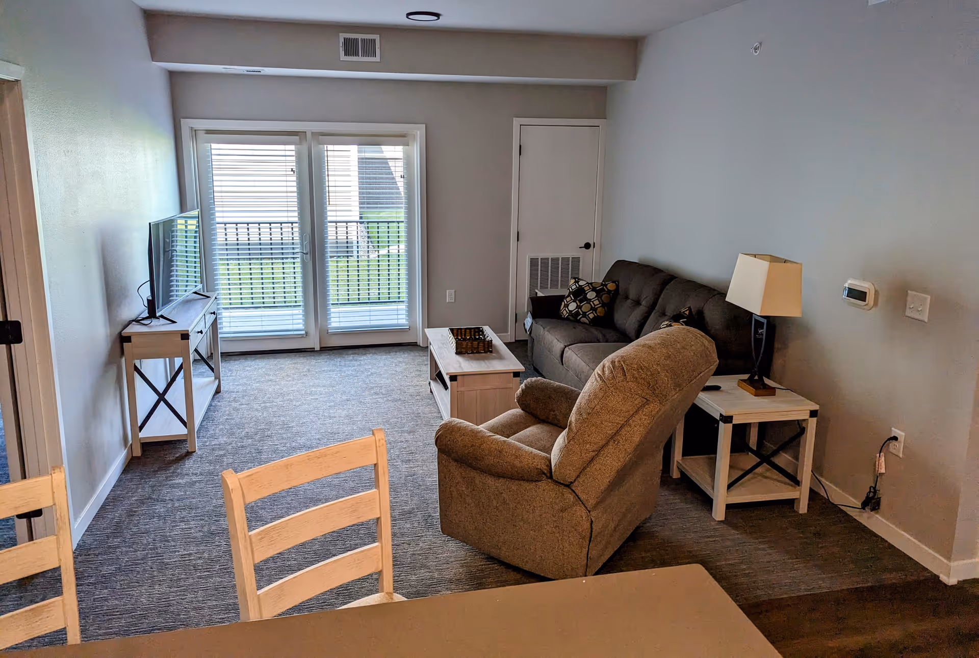 A cozy living room with a gray sofa, a beige recliner chair, a wooden coffee table, and a side table with a lamp. There is a TV on a stand against the left wall and glass doors with blinds leading outside. The room has neutral-colored walls and carpeted flooring.