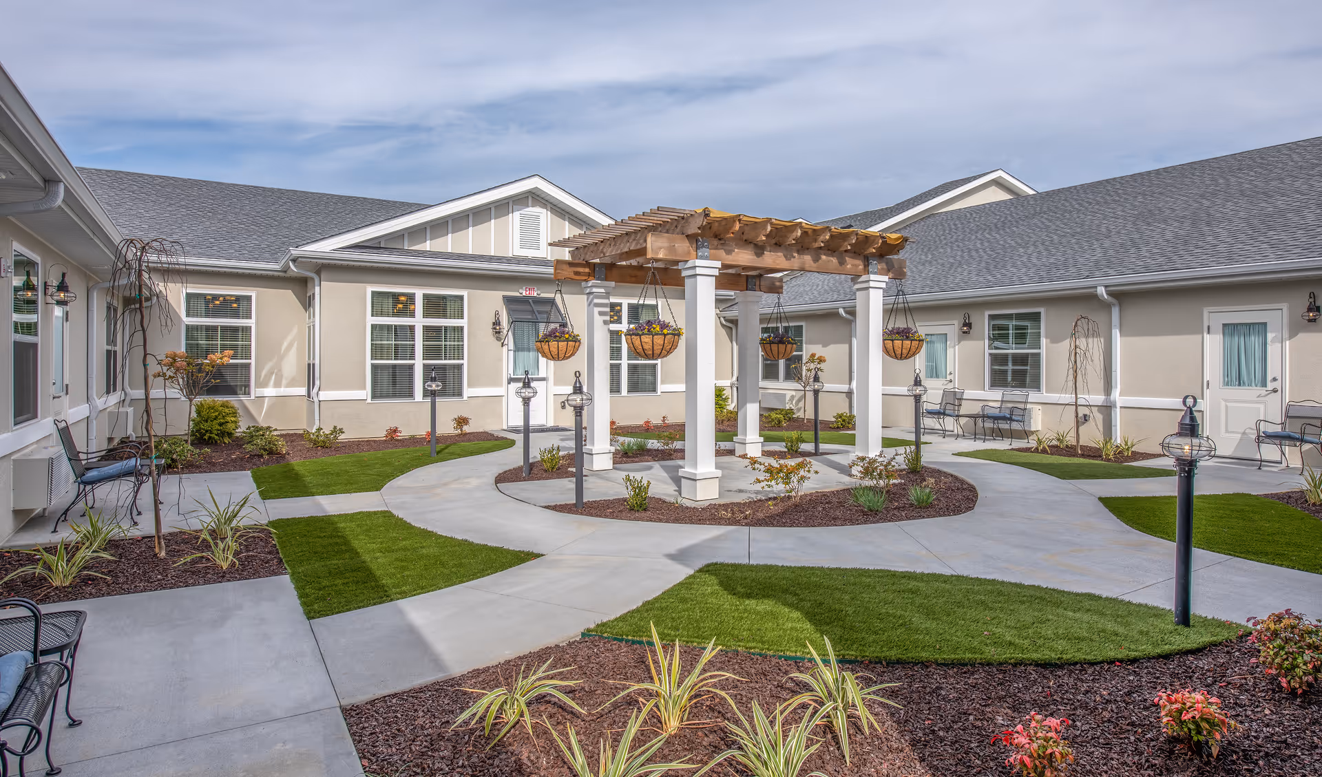 Sunny landscaped courtyard featuring a central pergola with hanging planters, paved walkways, benches, and surrounding single-story residential units.