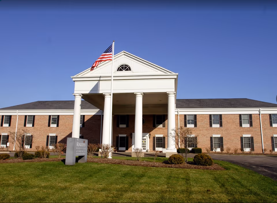 Front exterior view of Kingston Residence of Perrysburg, a two-story brick building with white columns at the entrance, an American flag on a flagpole, and a well-maintained lawn with shrubs.