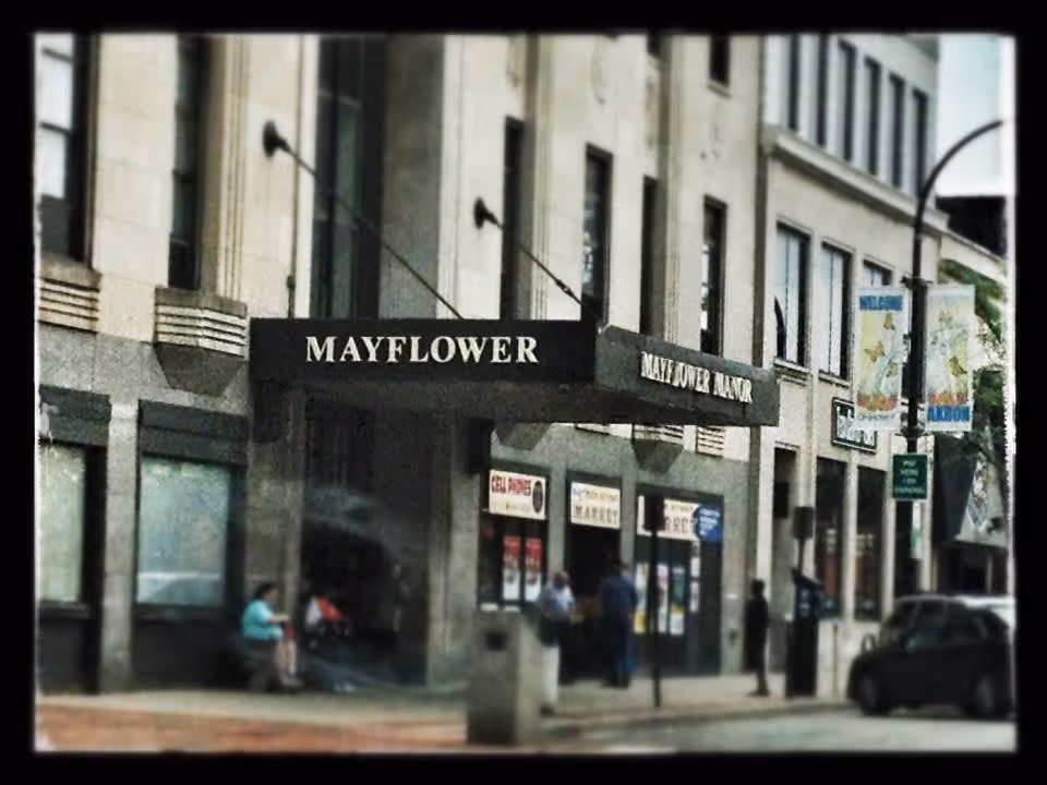 Street view of the entrance to Mayflower Manor Apartments, showing a building facade with a black awning displaying the name 'Mayflower Manor'. People are sitting and standing near the entrance, and there are cars parked along the street.