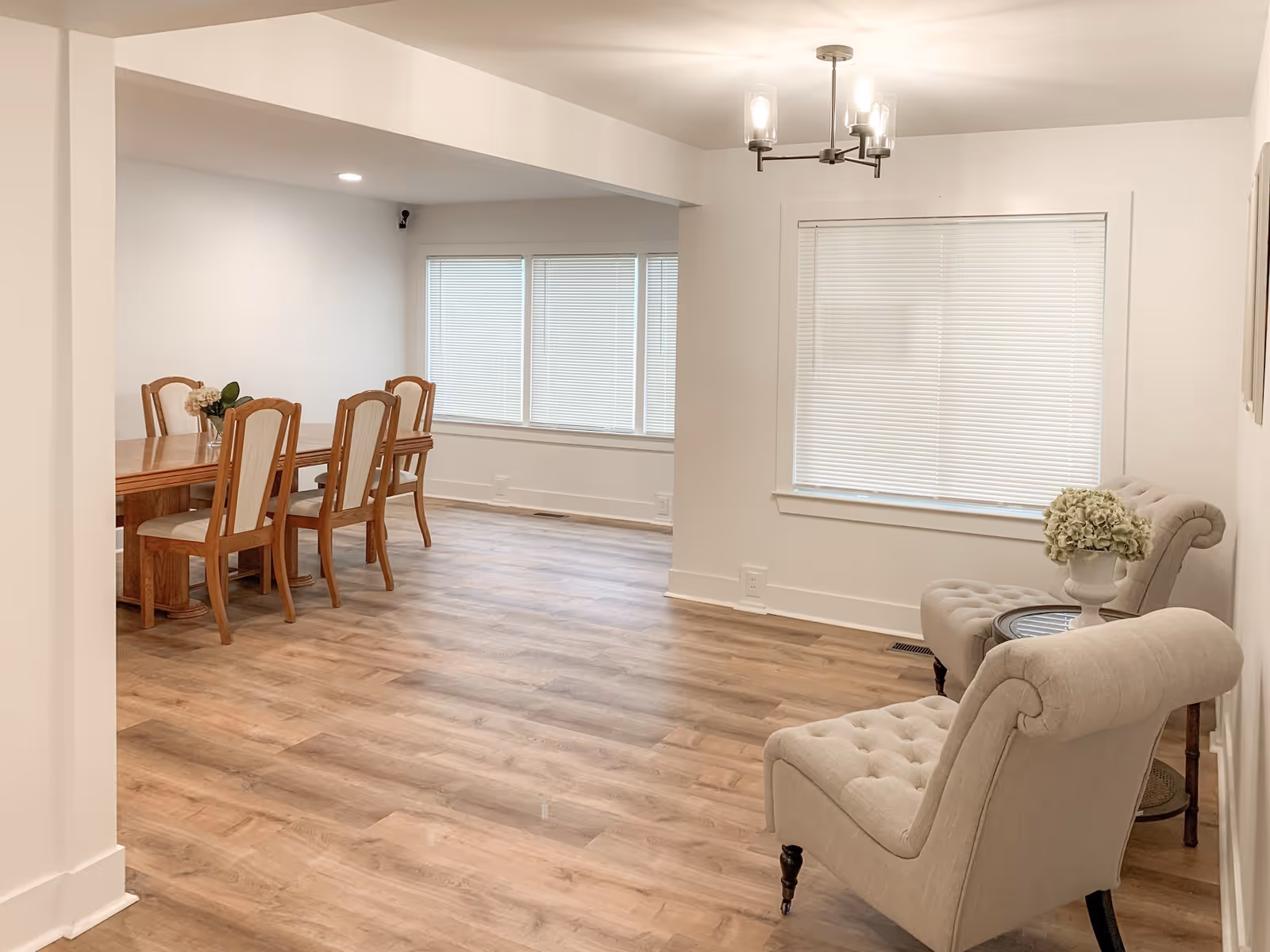 A bright and spacious interior room with light wood flooring and white walls. On the right side, there are two beige tufted armchairs with a small round table between them holding a vase of flowers. On the left side, there is a wooden dining table with six matching chairs. Large windows with white blinds allow natural light into the room. A modern ceiling light fixture is visible above.