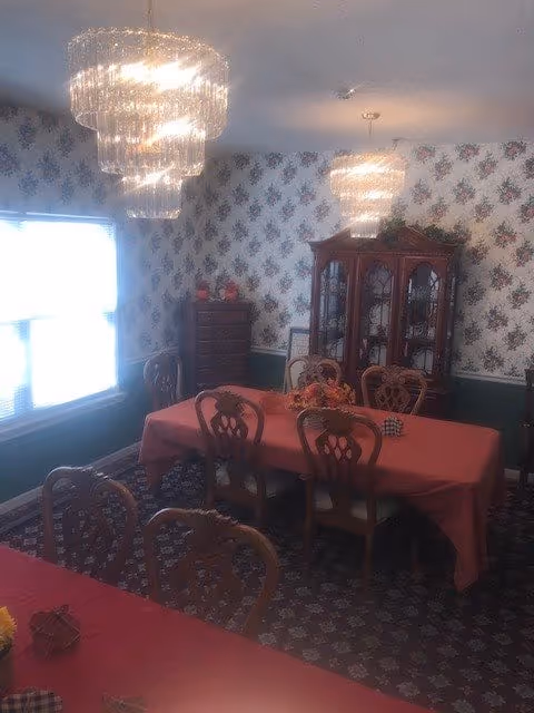 A traditional dining room with a long table covered in a red tablecloth surrounded by wooden chairs, a china cabinet against floral wallpaper, and two crystal chandeliers.