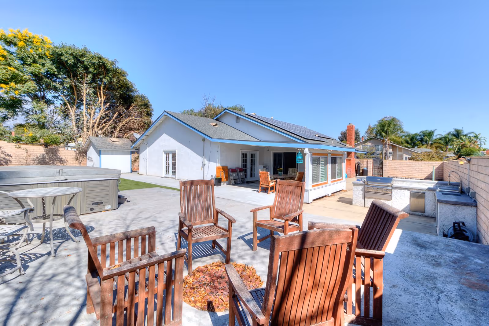 Outdoor patio area with wooden chairs arranged around a fire pit, a round metal table with chairs, a hot tub, and a built-in outdoor kitchen with a grill and sink. The area is enclosed by a brick wall and surrounded by trees and neighboring houses under a clear blue sky.