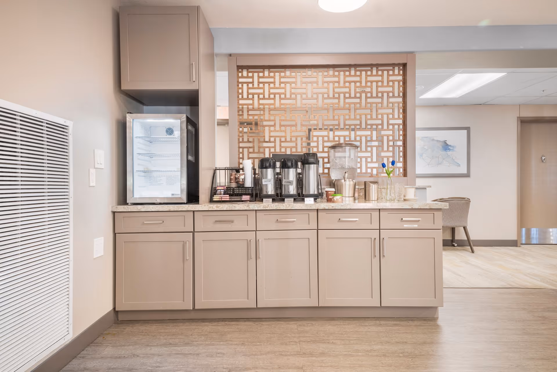 A coffee and beverage station in a senior living facility with beige cabinets, a small refrigerator, coffee dispensers, a water dispenser, cups, and a decorative wooden lattice panel behind the counter. The area is clean and well-lit with neutral tones and a glimpse of a seating area in the background.