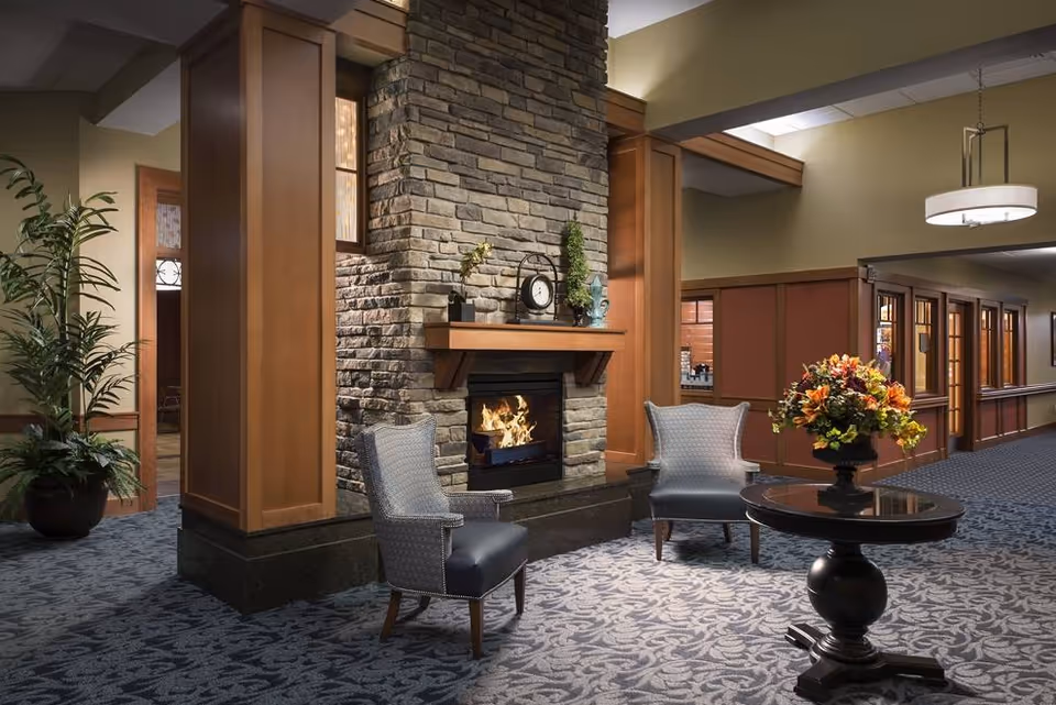 A cozy interior lounge area with a stone fireplace in the center, two upholstered armchairs positioned in front of the fireplace, a round wooden table with a floral arrangement, and wooden paneling on the walls. The space is carpeted with a patterned design and features warm lighting.