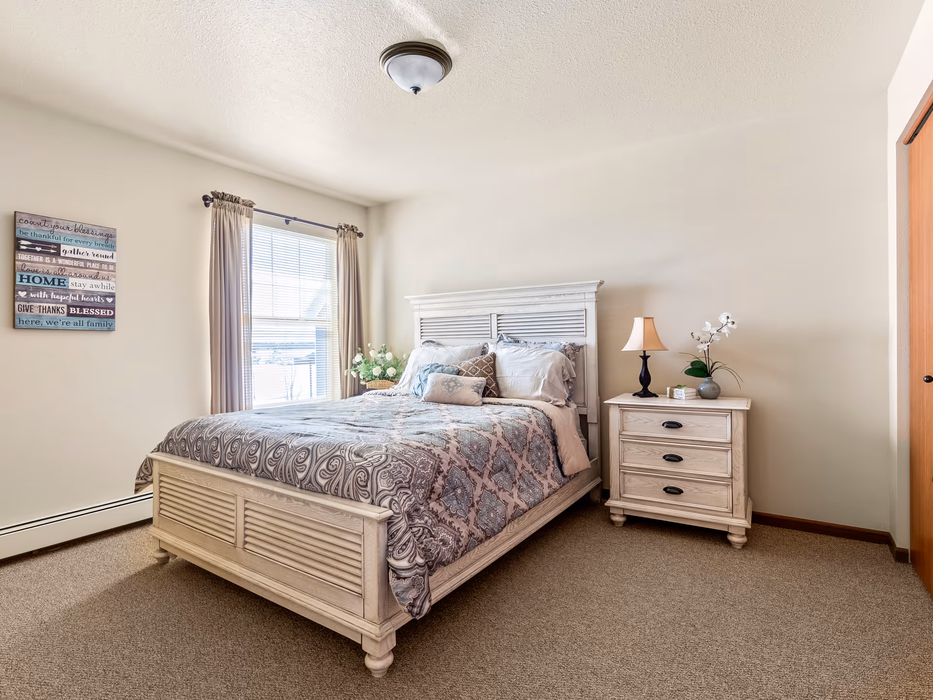 A bright bedroom with a large bed featuring a patterned comforter and multiple pillows. Next to the bed is a wooden nightstand with a lamp and a small plant. A window with beige curtains allows natural light to fill the room. A decorative sign with inspirational text hangs on the wall beside the window.