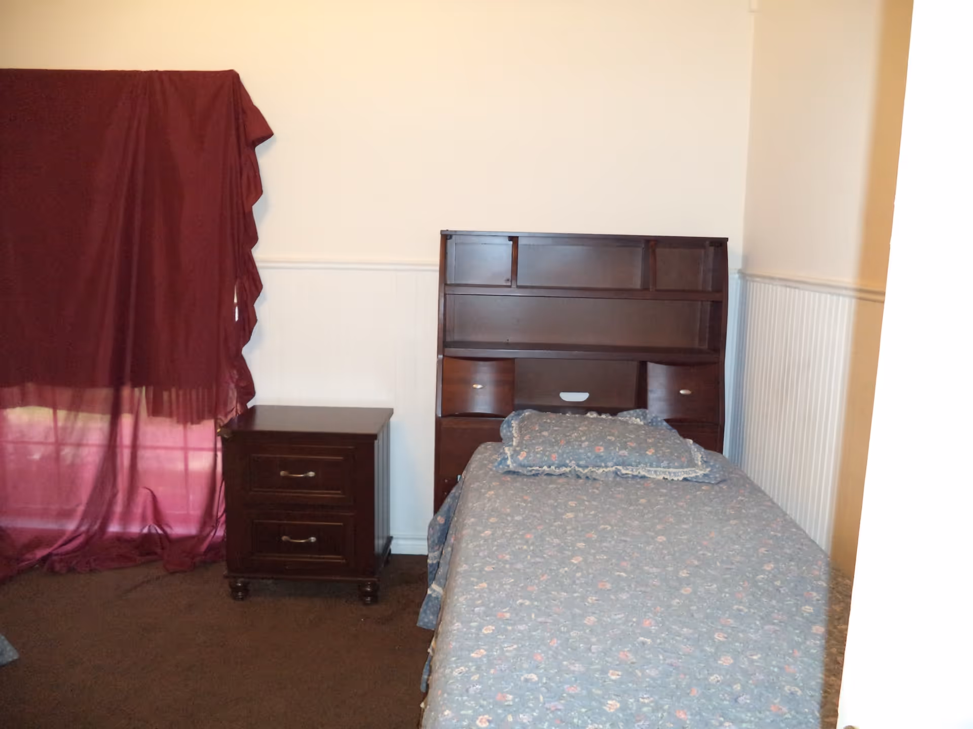 A small bedroom with a single bed covered in a blue floral bedspread and matching pillow. Next to the bed is a dark wooden nightstand with two drawers. A window with a maroon curtain is partially visible on the left side of the room. The walls are painted white with a white wainscoting panel halfway up.