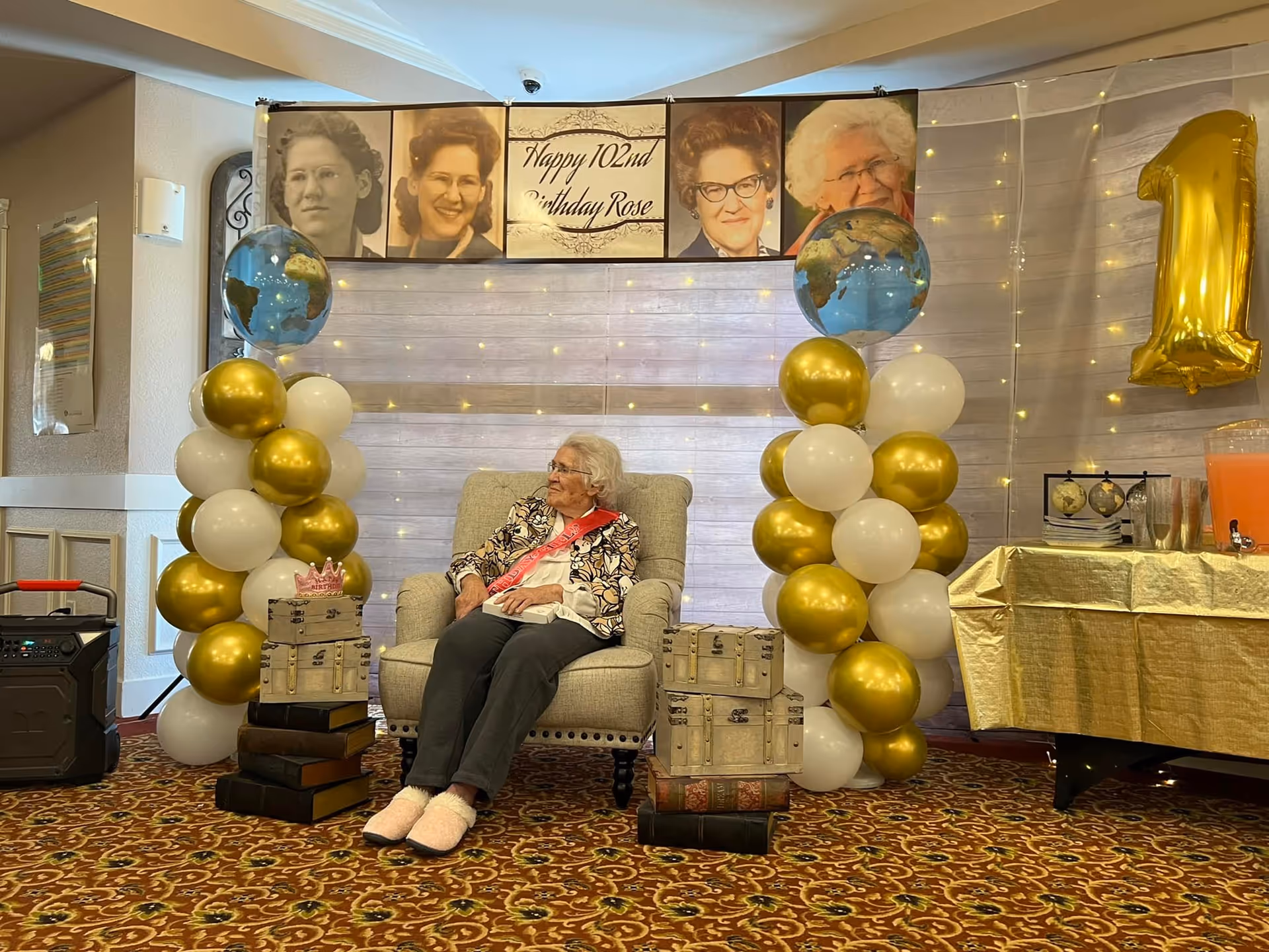 An elderly woman wearing a floral jacket and a pink sash sits in a beige armchair in a decorated room. Behind her are two columns of gold and white balloons topped with globe balloons. Above her is a banner with four portraits of the same woman at different ages and the text 'Happy 102nd Birthday Rose'. To the right is a table covered with a gold tablecloth and a large gold balloon shaped like the number 1. The floor has a patterned carpet.