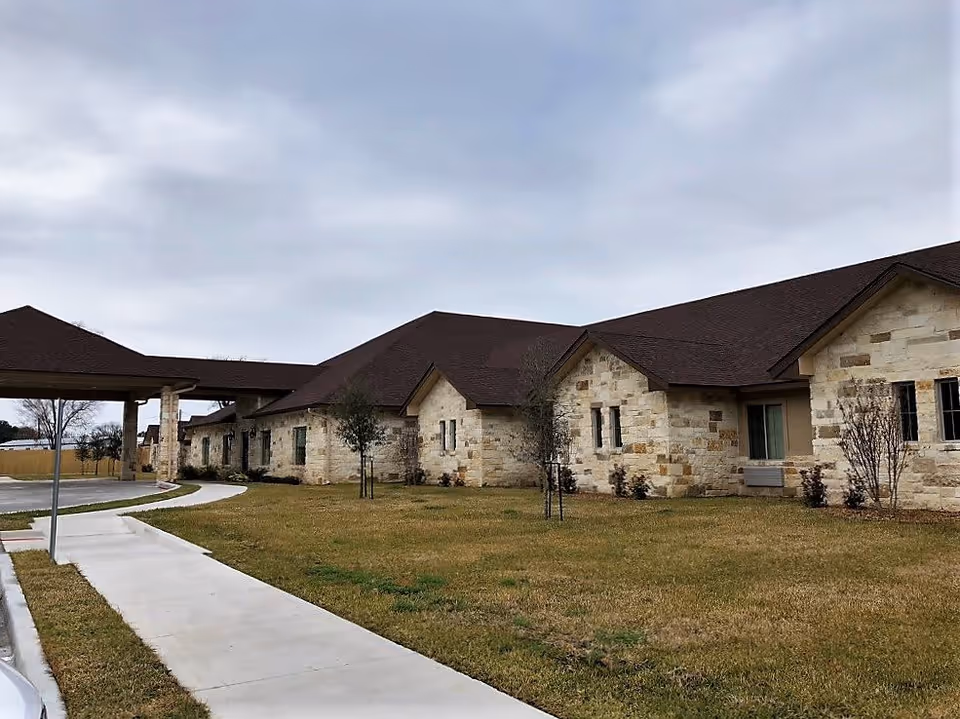 Exterior view of a single-story building with stone walls and a dark brown roof, surrounded by a grassy lawn and a sidewalk leading to a covered entrance area under a cloudy sky.
