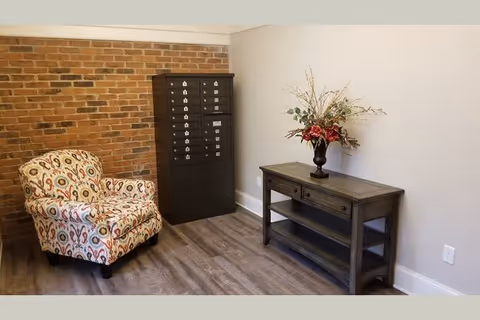 A small interior room with a patterned armchair, a black mailbox unit against a brick wall, and a dark wooden console table with a floral arrangement on top, set against a plain light-colored wall and wood flooring.