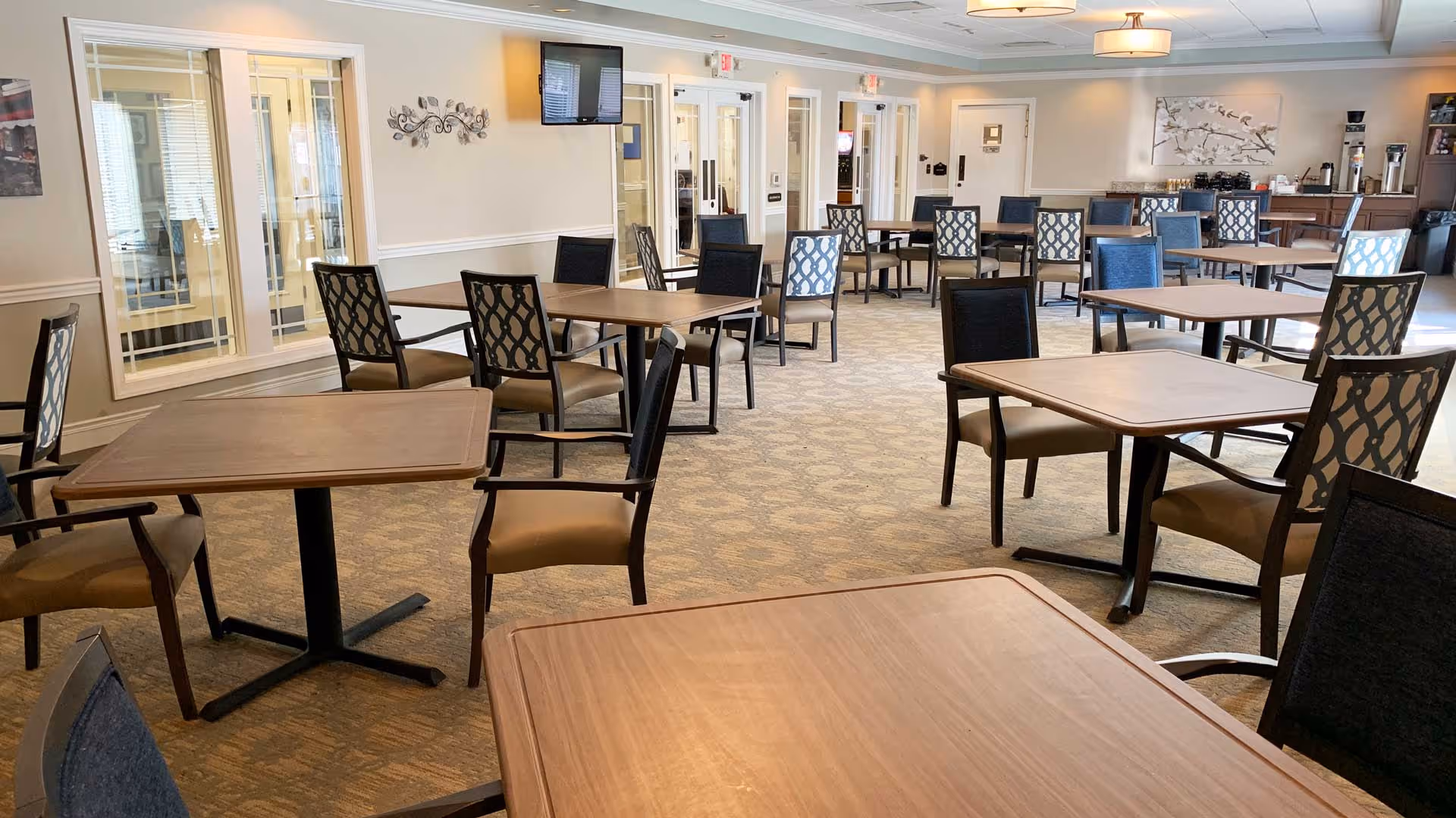 Spacious dining room with multiple wooden tables and upholstered chairs, a wall-mounted TV, and a beverage station in the background.