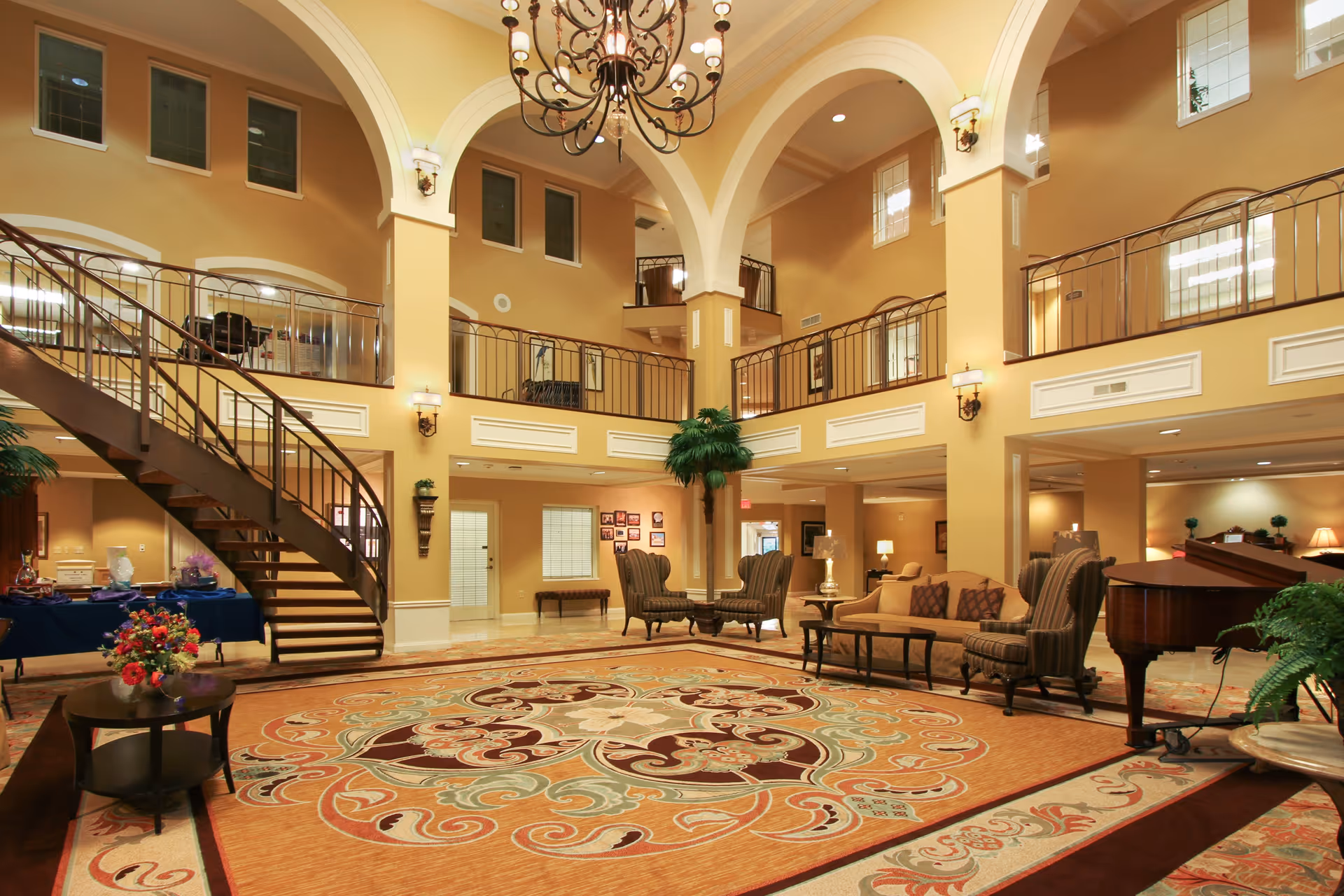 Spacious two-story lobby with a sweeping curved staircase, chandelier, seating areas, and an ornate patterned rug.