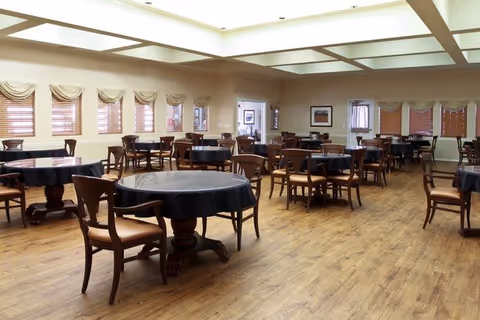 A spacious dining room with multiple round tables covered with black tablecloths, each surrounded by wooden chairs with cushioned seats. The room has large windows with blinds and valances, light-colored walls, and a ceiling with recessed lighting panels. The floor is made of wood.
