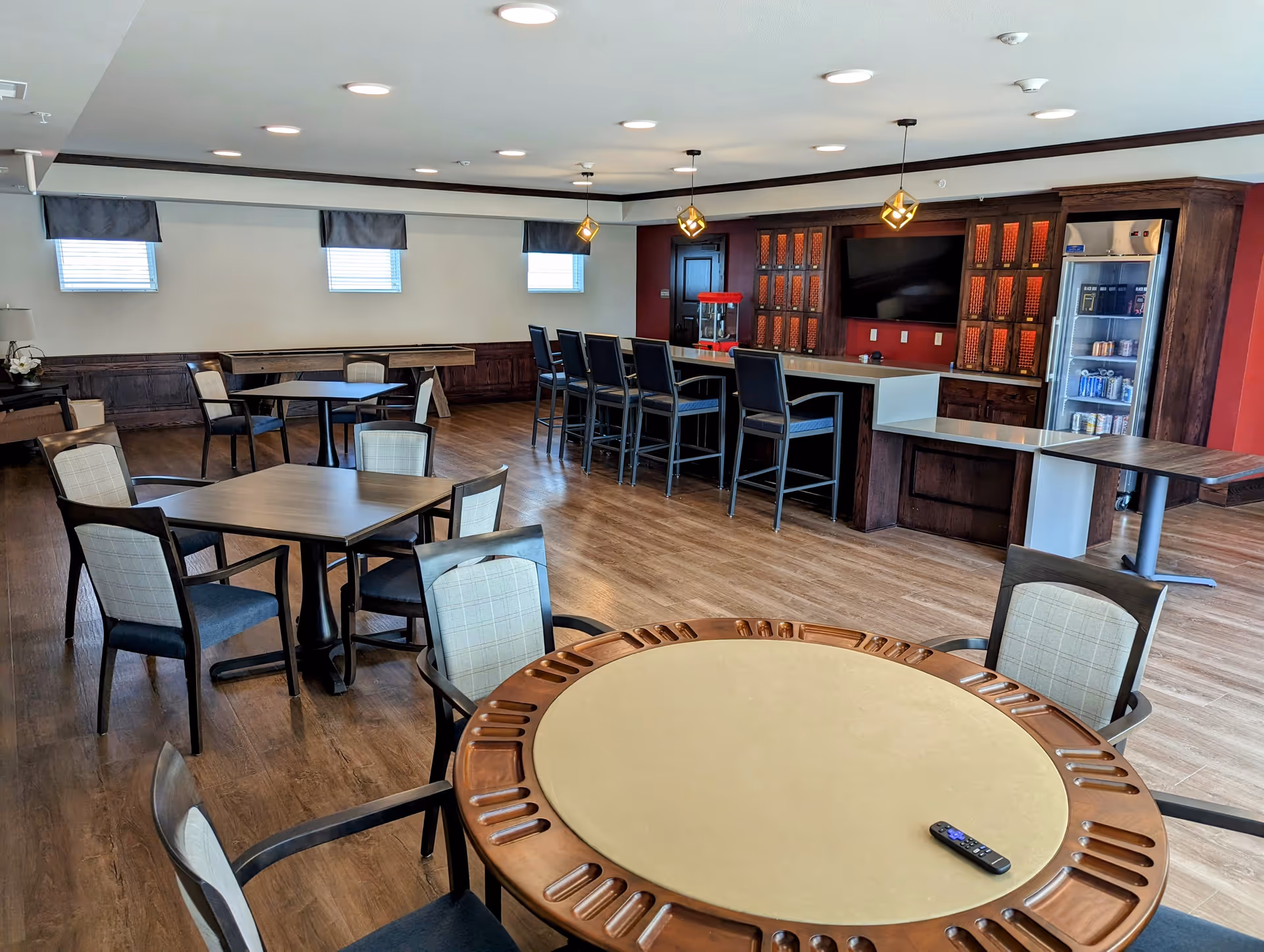 A spacious common area with multiple tables and chairs, including a round table with a remote control on it in the foreground. There is a bar counter with high chairs, a popcorn machine, a large flat-screen TV mounted on a wooden cabinet, and a refrigerator stocked with beverages. The room has wooden flooring, several small windows with valances, and ceiling lights.