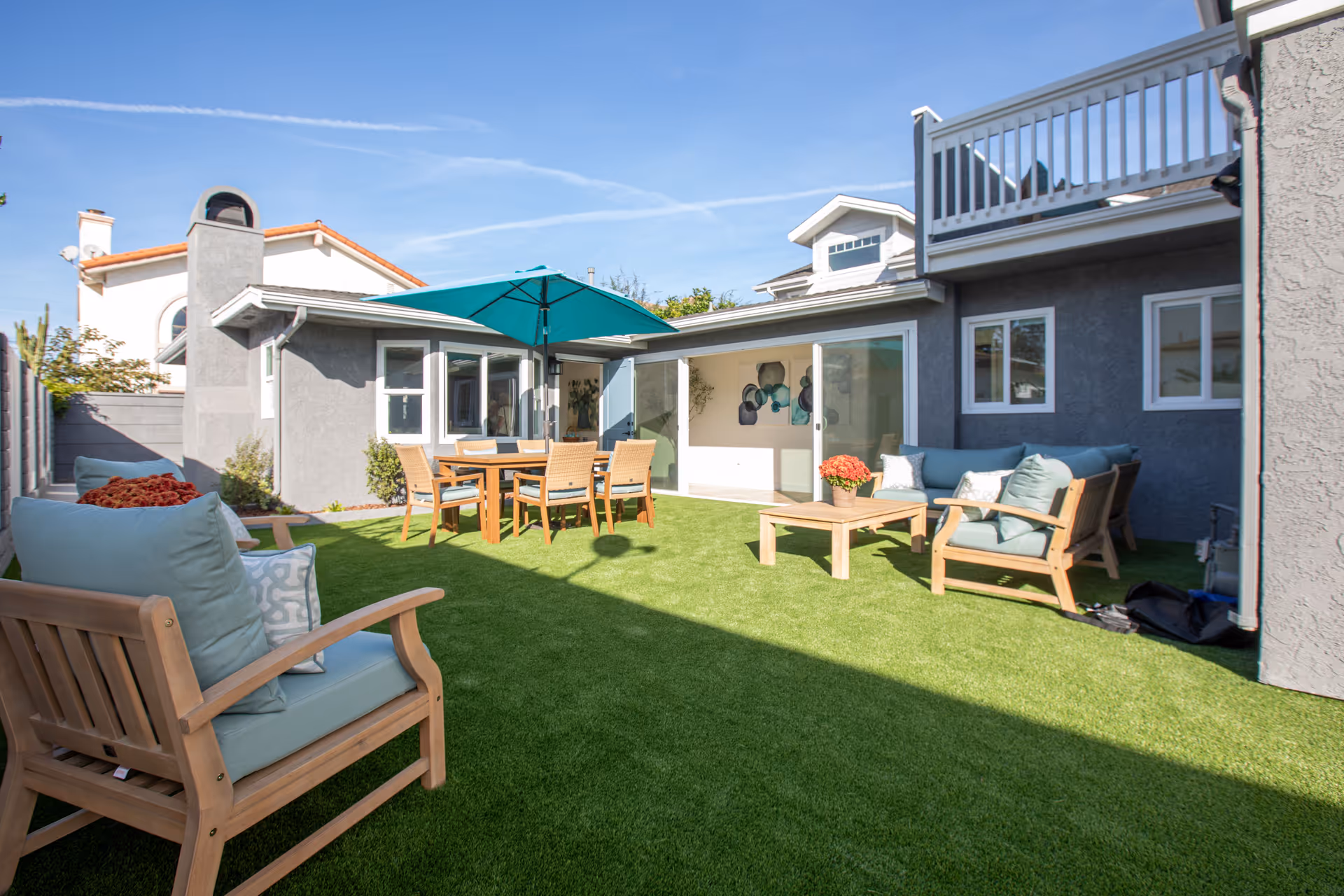 Outdoor patio area with artificial grass, wooden seating furniture with light blue cushions, a wooden dining table with chairs under a teal umbrella, and a gray building with sliding glass doors and windows under a clear blue sky.