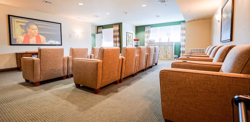 A cozy media room with two rows of brown upholstered armchairs facing a large wall-mounted television displaying a woman in a red dress. The room has beige walls, carpeted floor, and windows with striped curtains allowing natural light to enter. There is a popcorn machine and decorative items on a cabinet near the windows.