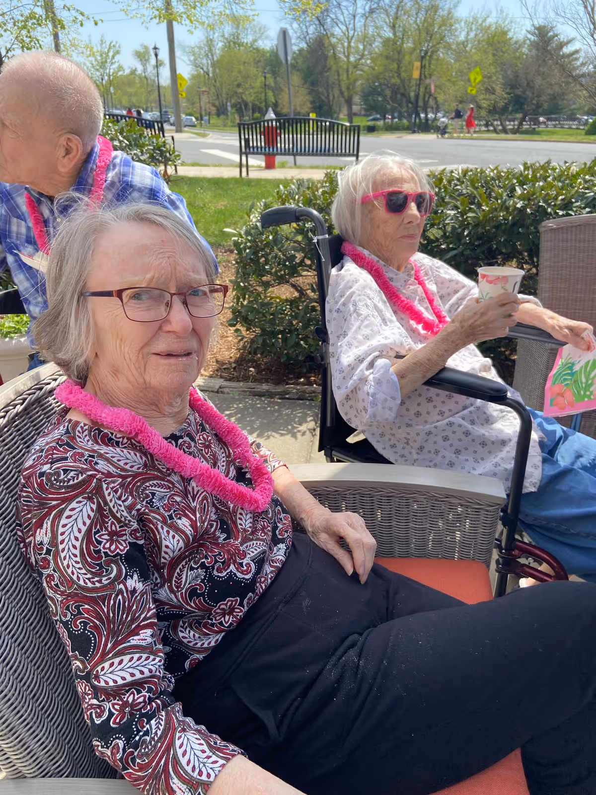 Two elderly women sitting outdoors on wicker chairs. One woman is wearing glasses and a patterned top with a pink lei around her neck, while the other woman is in a wheelchair wearing pink sunglasses, a white patterned blouse, and a pink lei, holding a paper cup and a colorful card. A man in a blue plaid shirt with a pink lei is partially visible in the background. The setting is a sunny day with greenery and a street visible behind them.