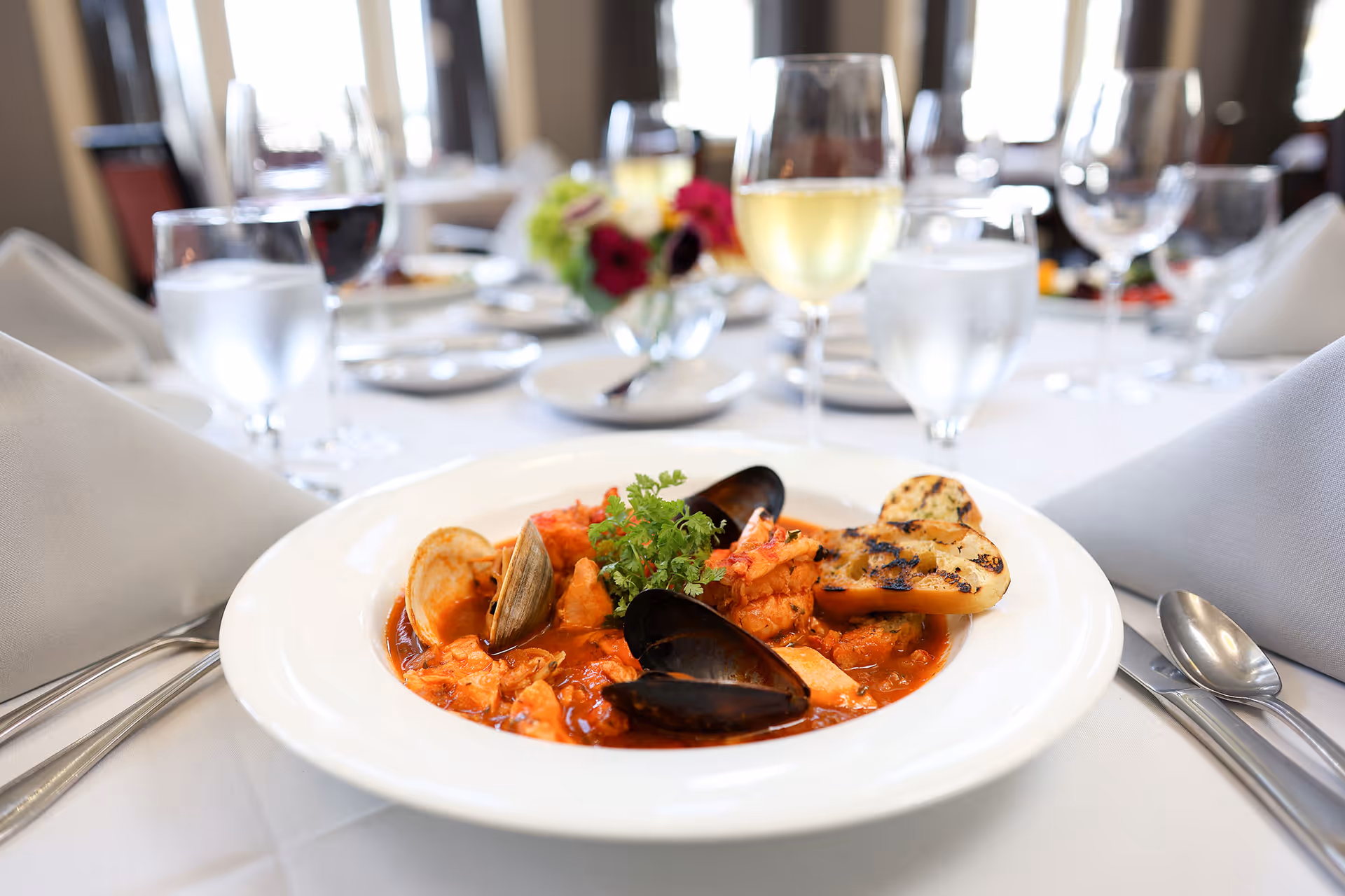 A plated seafood stew with mussels and grilled bread on a white tablecloth in a formal dining setting with wine glasses.