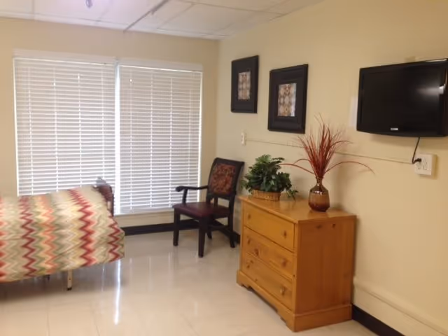 A simple resident bedroom with a bed, chair, wooden dresser topped with plants and a vase, a wall-mounted TV, and large window blinds.