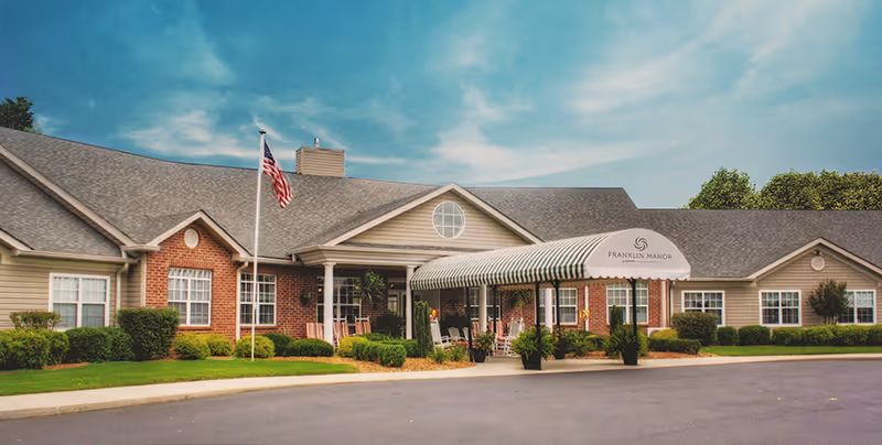 Exterior view of Franklin Manor, a single-story senior living facility with a brick and siding facade, a covered entrance with striped awning, an American flag on a flagpole, and well-maintained landscaping under a partly cloudy sky.