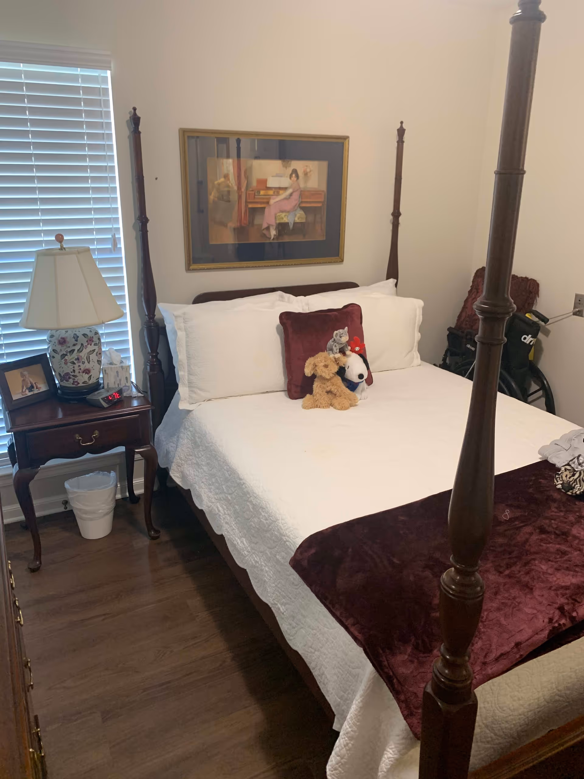 A cozy bedroom with a four-poster wooden bed dressed in white bedding and a burgundy throw blanket. On the bed are three stuffed animals and a burgundy pillow. To the left of the bed is a wooden nightstand with a floral lamp, a framed photo, a clock, and a tissue box. A window with white blinds is behind the nightstand. A framed painting of a woman playing piano hangs on the wall above the bed. The floor is wooden, and a wheelchair is partially visible in the corner.