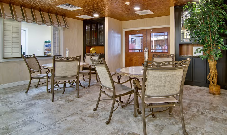 Small communal dining area with round tables and metal-framed chairs, tile floor, and a service window.