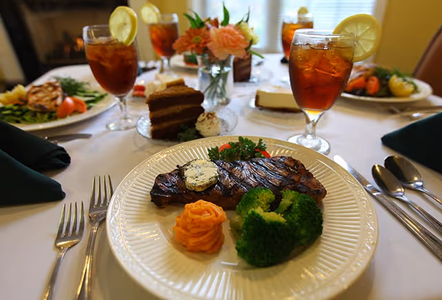 A plated grilled steak with herbed butter, broccoli, mashed sweet potato, iced tea and desserts arranged on a white-clothed dining table with a floral centerpiece.