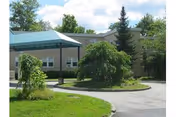 Entrance of a senior living residence with a covered porte-cochère, circular driveway, and landscaped lawn and trees.
