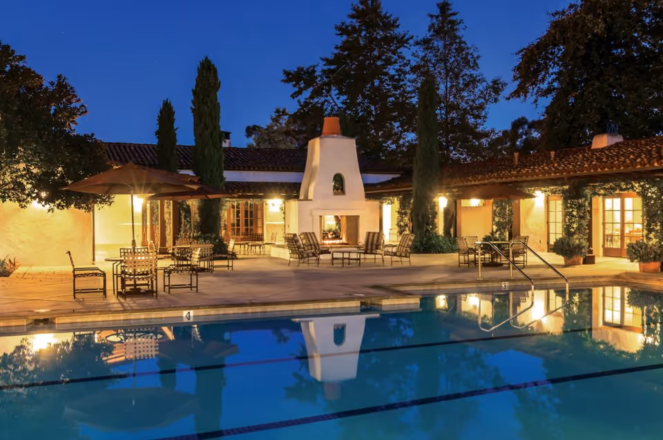 Outdoor patio area at dusk with a swimming pool in the foreground, surrounded by chairs and tables with umbrellas. There is a large white outdoor fireplace in the center, tall trees, and a building with warm lights and tiled roof in the background.