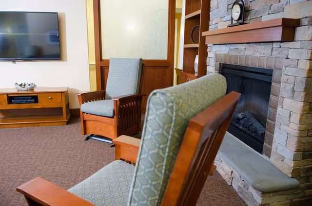 A cozy living room area featuring two wooden armchairs with patterned cushions, a stone fireplace with a clock on the mantel, a wooden TV stand with a flat-screen television mounted on the wall, and a carpeted floor.