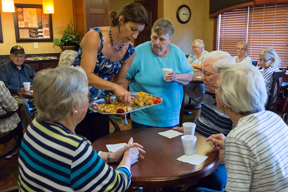 A group of elderly people sitting around tables in a communal dining area, being served food by a woman holding a tray with various dishes. The room has warm lighting, wooden blinds, and framed artwork on the walls.