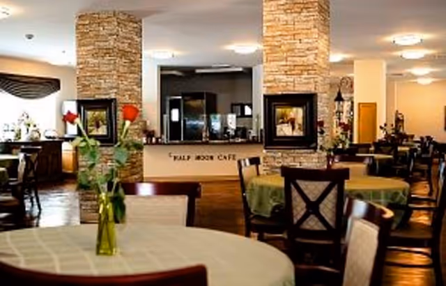 Interior view of a dining area with round tables covered with tablecloths and chairs around them. A vase with red roses is placed on the table in the foreground. The background shows stone pillars, framed pictures, and a counter labeled 'Half Moon Cafe'.
