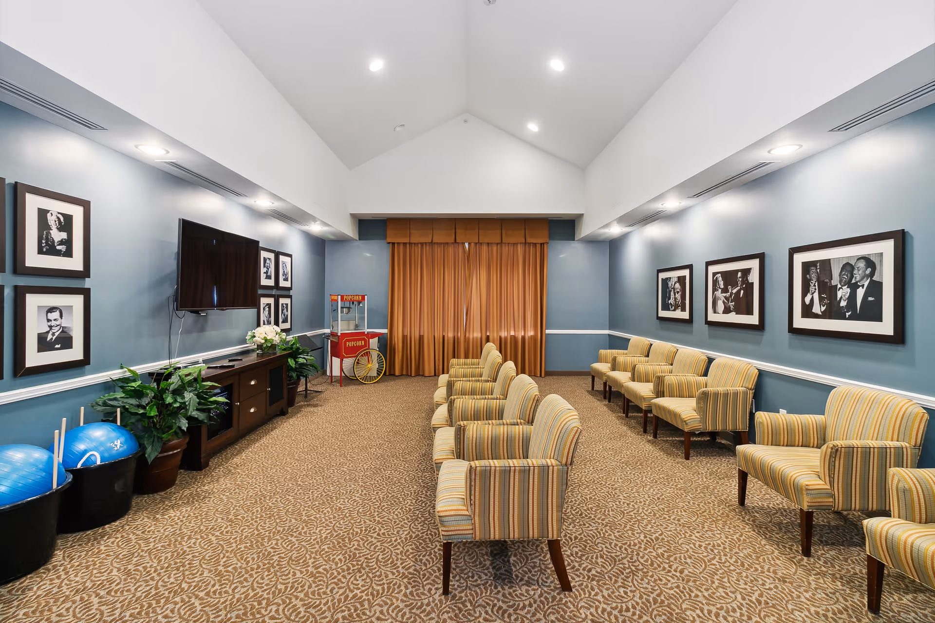 A community room with rows of striped armchairs facing a wall-mounted flat screen TV. The walls are painted blue and decorated with black and white framed photographs. There is a popcorn machine in the corner and two large blue exercise balls in black bins near a wooden cabinet with plants on top. The room has a high vaulted ceiling with recessed lighting and a patterned carpet.