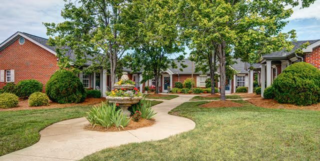 A landscaped outdoor courtyard area at Brekenridge Retirement Center featuring a winding concrete pathway, a tiered stone fountain with flowers, green grass, bushes, and several trees. The courtyard is surrounded by single-story red brick buildings with white trim and windows.