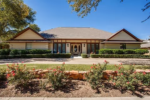 Front exterior view of a single-story building with a beige brick facade, a gray shingled roof, and symmetrical design. The entrance features double doors with glass panels, flanked by large windows. There are neatly trimmed bushes along the front of the building and a flower bed with red flowers and a low stone border in the foreground. Trees with green leaves are visible on either side of the building under a clear blue sky.
