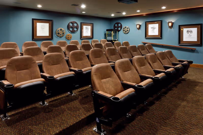 A small theater room with three rows of brown cushioned seats arranged in a tiered layout. The walls are painted teal and decorated with framed black and white portraits and vintage film reels. A popcorn machine is visible in the back corner of the room.