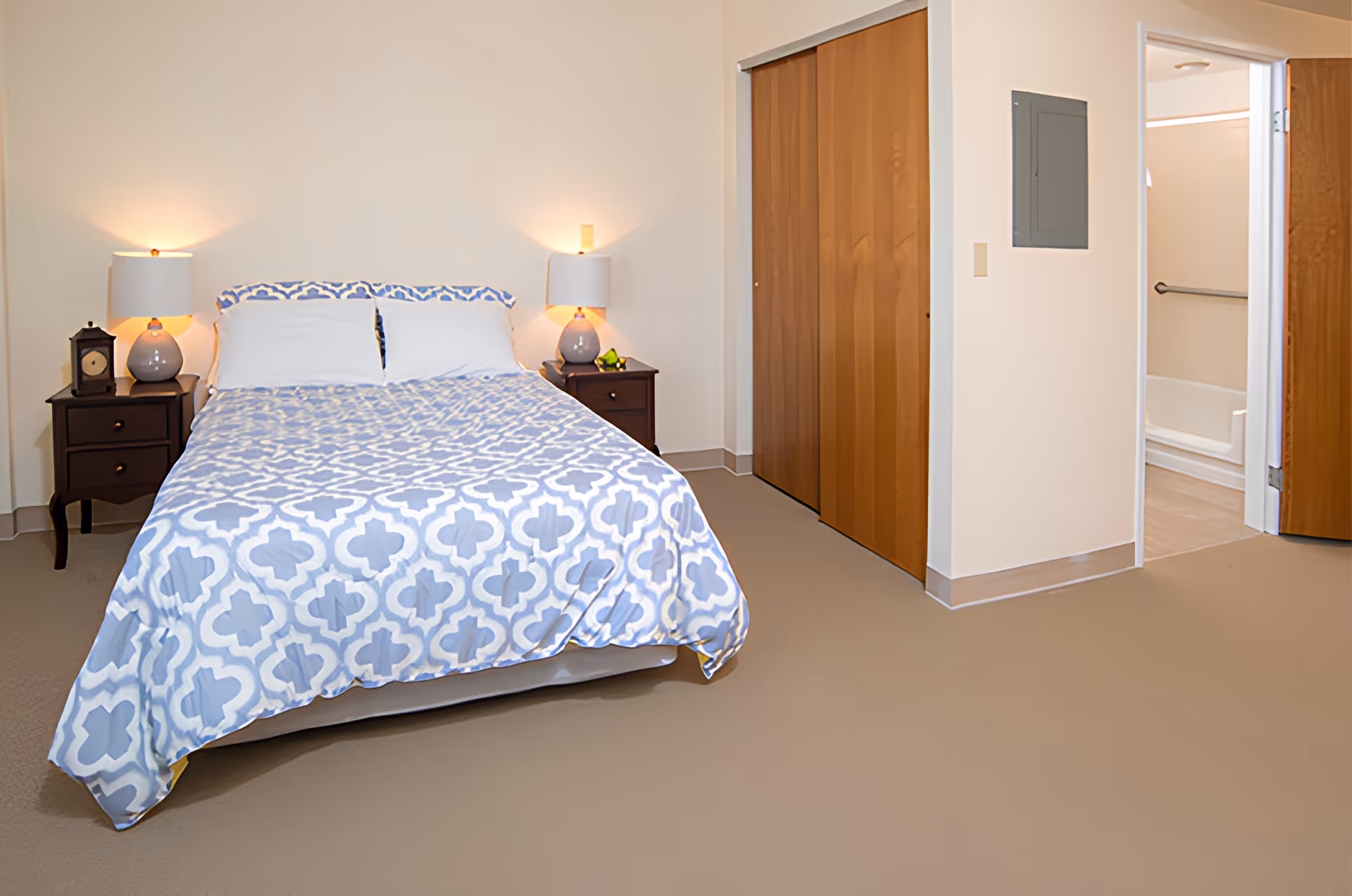 A bedroom in Western Hills Retirement Village featuring a bed with a blue and white patterned comforter, two bedside tables each with a lamp, a clock on the left table, a wooden closet with sliding doors, and an open door leading to a bathroom with a bathtub and grab bar.