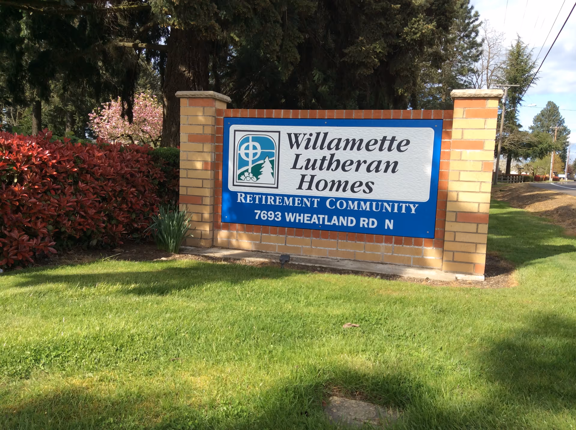 A brick sign for Willamette Lutheran Homes Retirement Community located at 7693 Wheatland Rd N, surrounded by green grass, red bushes, and trees in the background.