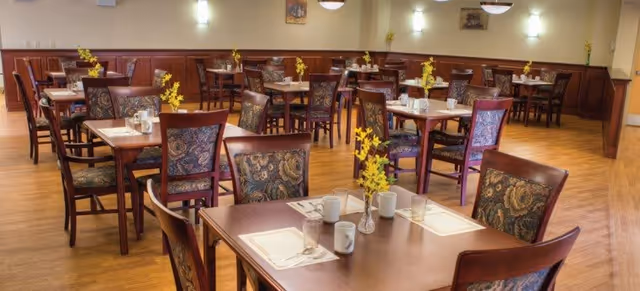 A dining room with multiple wooden tables and chairs arranged neatly. Each table is set with placemats, cups, glasses, and a small vase with yellow flowers. The room has wooden flooring and light-colored walls with wall-mounted lights and framed pictures.