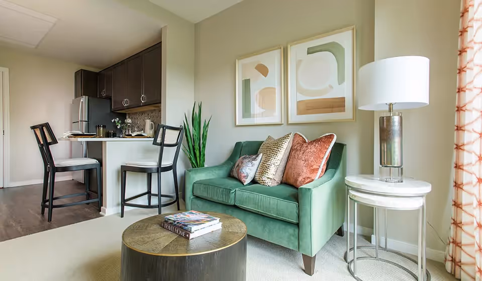 A cozy living area featuring a green loveseat with decorative pillows, a round wooden coffee table with books on it, a side table with a modern lamp, and two black chairs at a kitchen counter. The kitchen area includes dark cabinets and a stainless steel refrigerator. Two abstract art pieces hang on the wall above the loveseat.